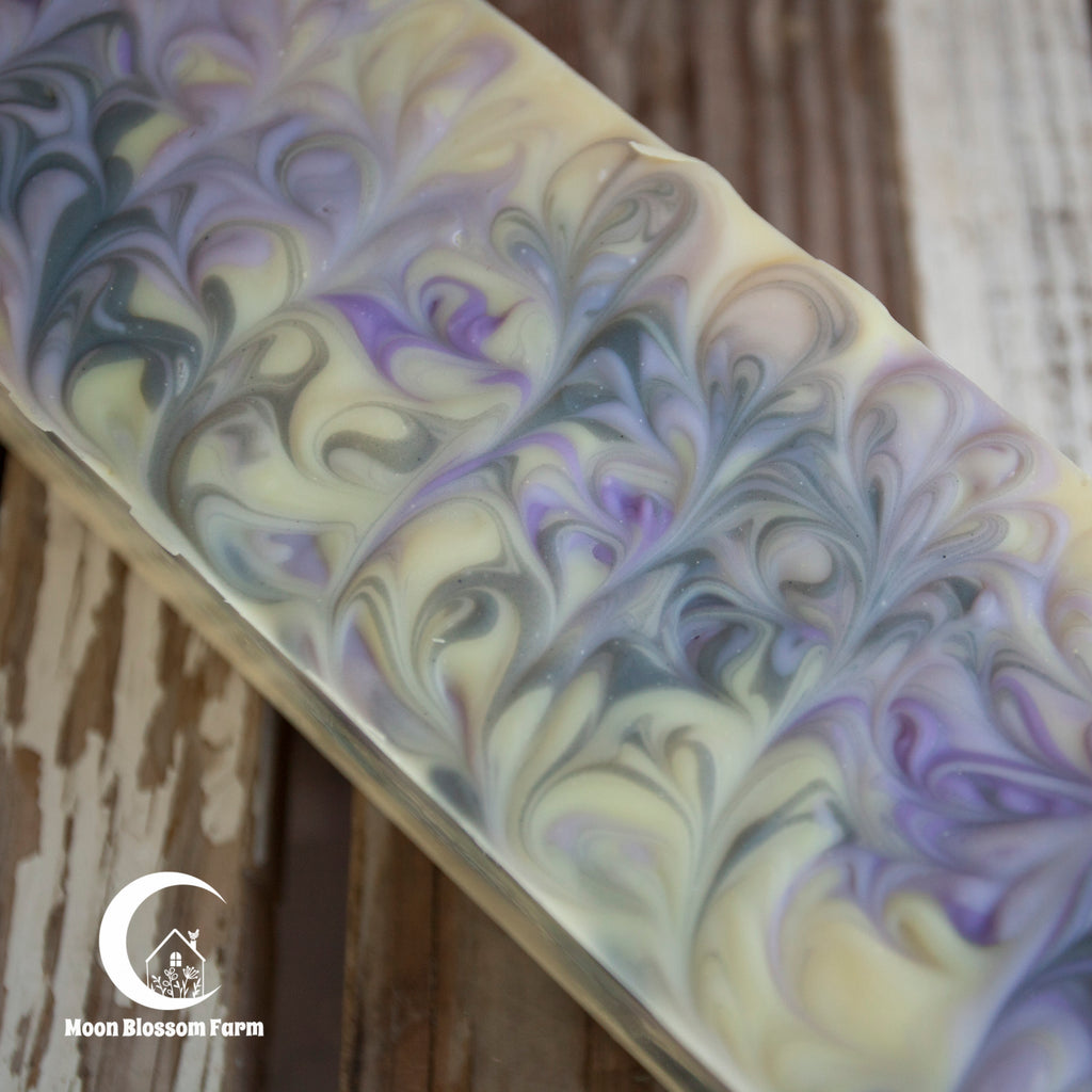 Marbled soap bar with purple and white swirls on a wooden background, featuring the Moon Blossom Farm logo.