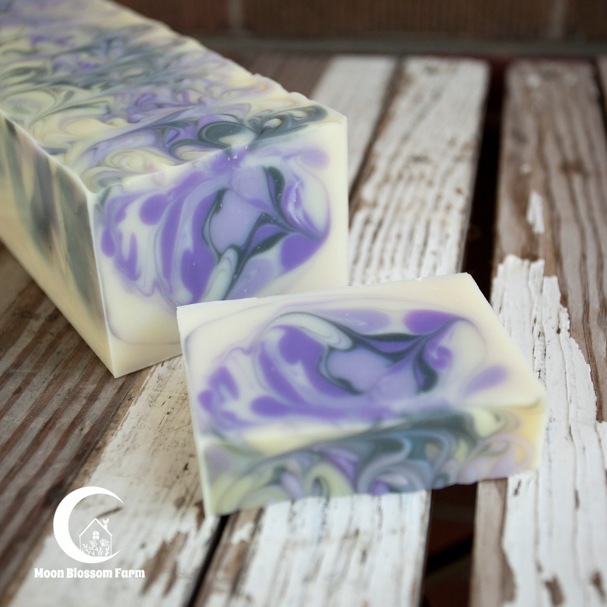 Two bars of soap with purple and black swirls on a wooden surface, branded 'Moon Blossom Farm'.