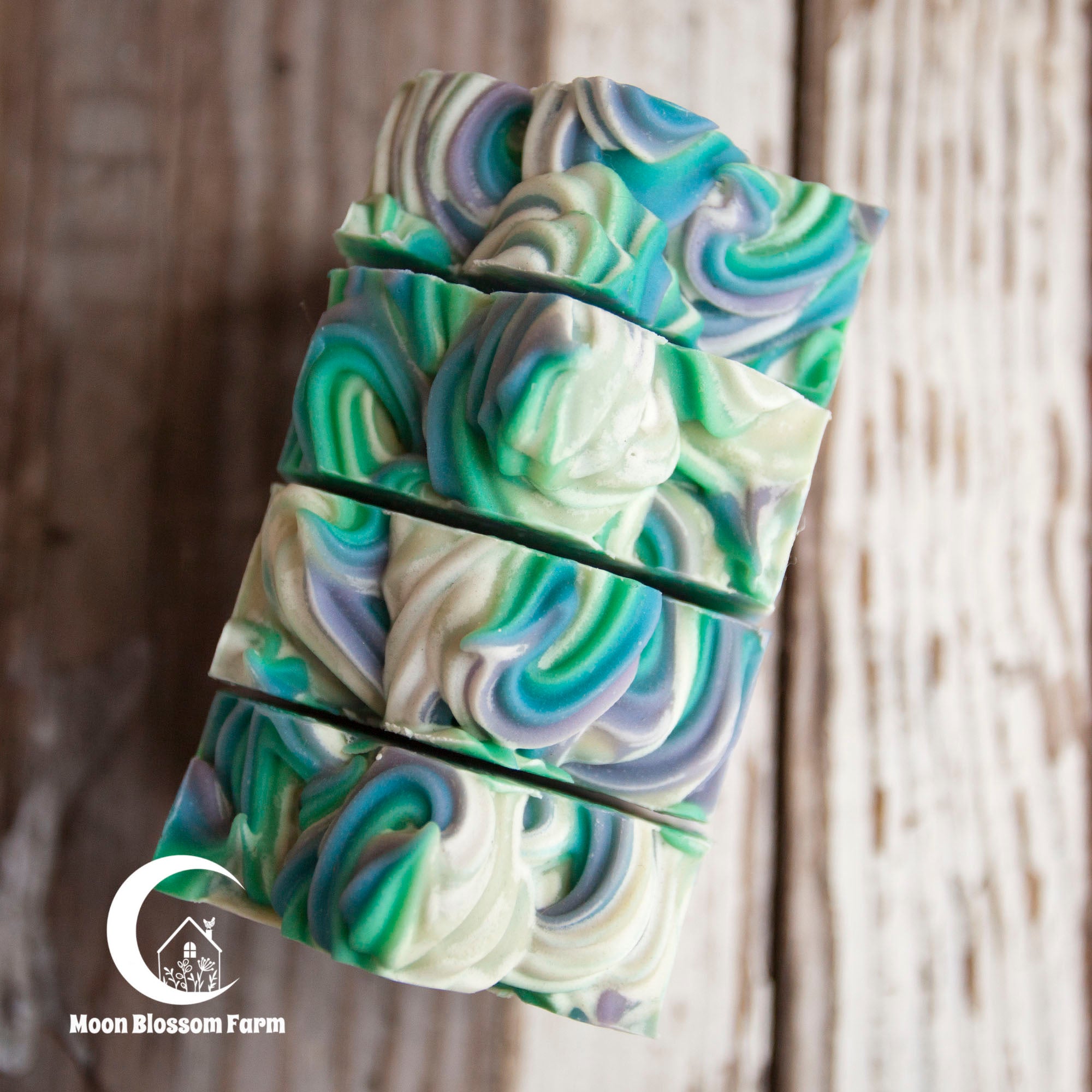 Swirled soap bar with green, blue, and white colors on a wooden background, featuring the Moon Blossom Farm logo.