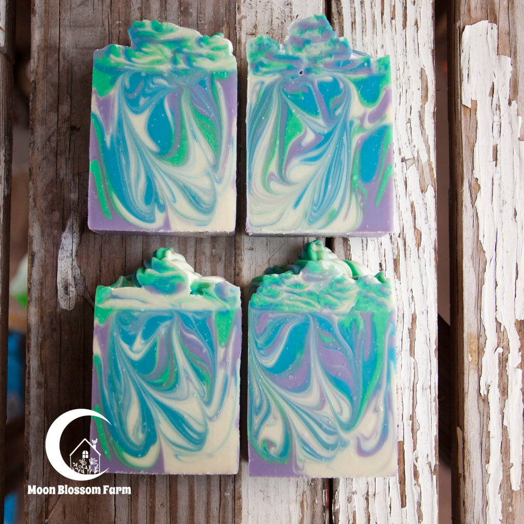 Four bars of marbled soap in green, blue, and purple on a wooden background with 'Moon Blossom Farm' logo.