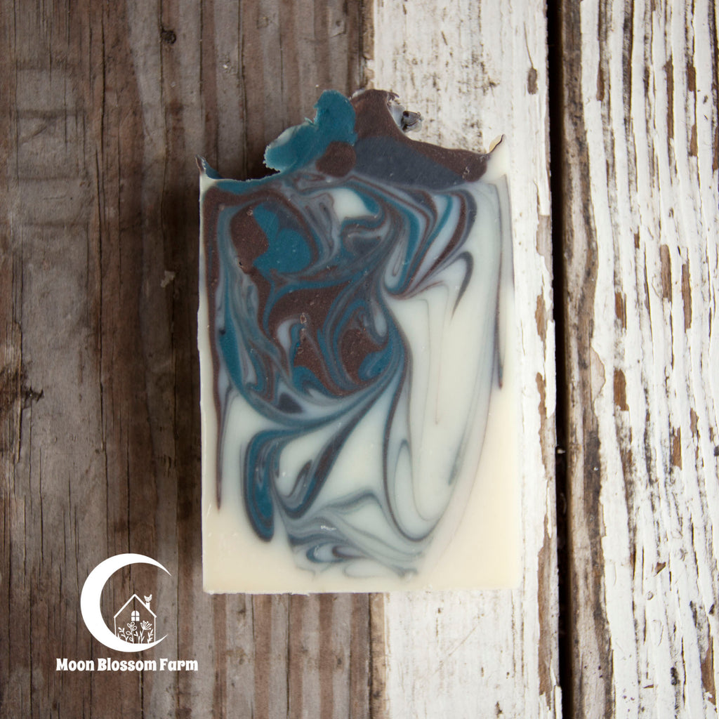 Marbled soap bar with brown and blue swirls on a wooden background, featuring Moon Blossom Farm logo.