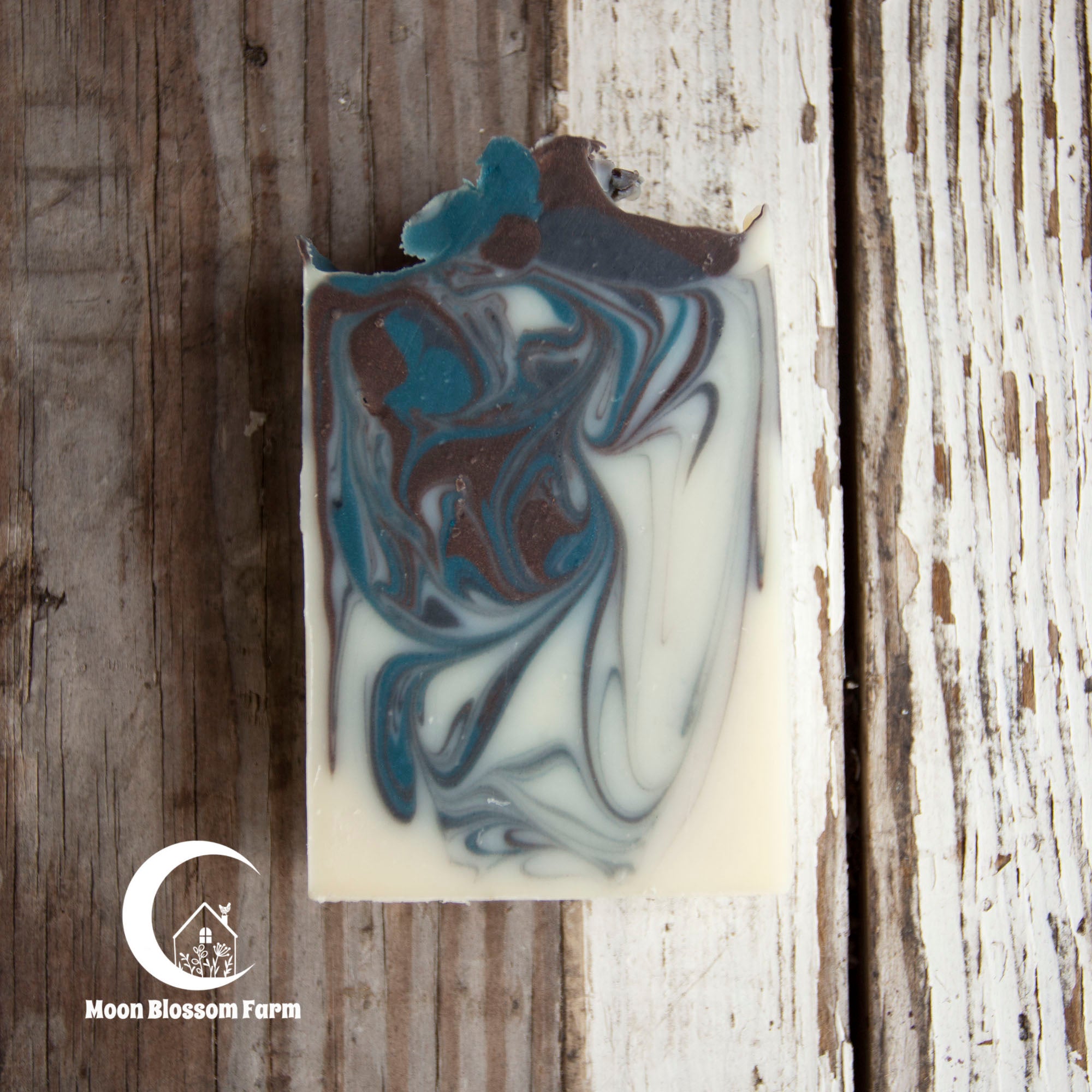 Marbled soap bar with brown and blue swirls on a wooden background, featuring Moon Blossom Farm logo.