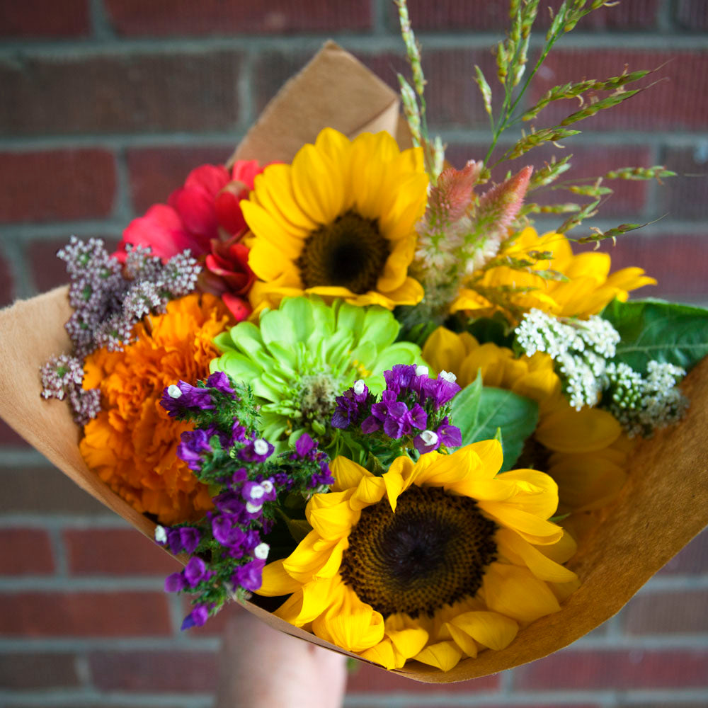 Bouquet of sunflowers and other flowers wrapped in brown paper against a brick wall.
